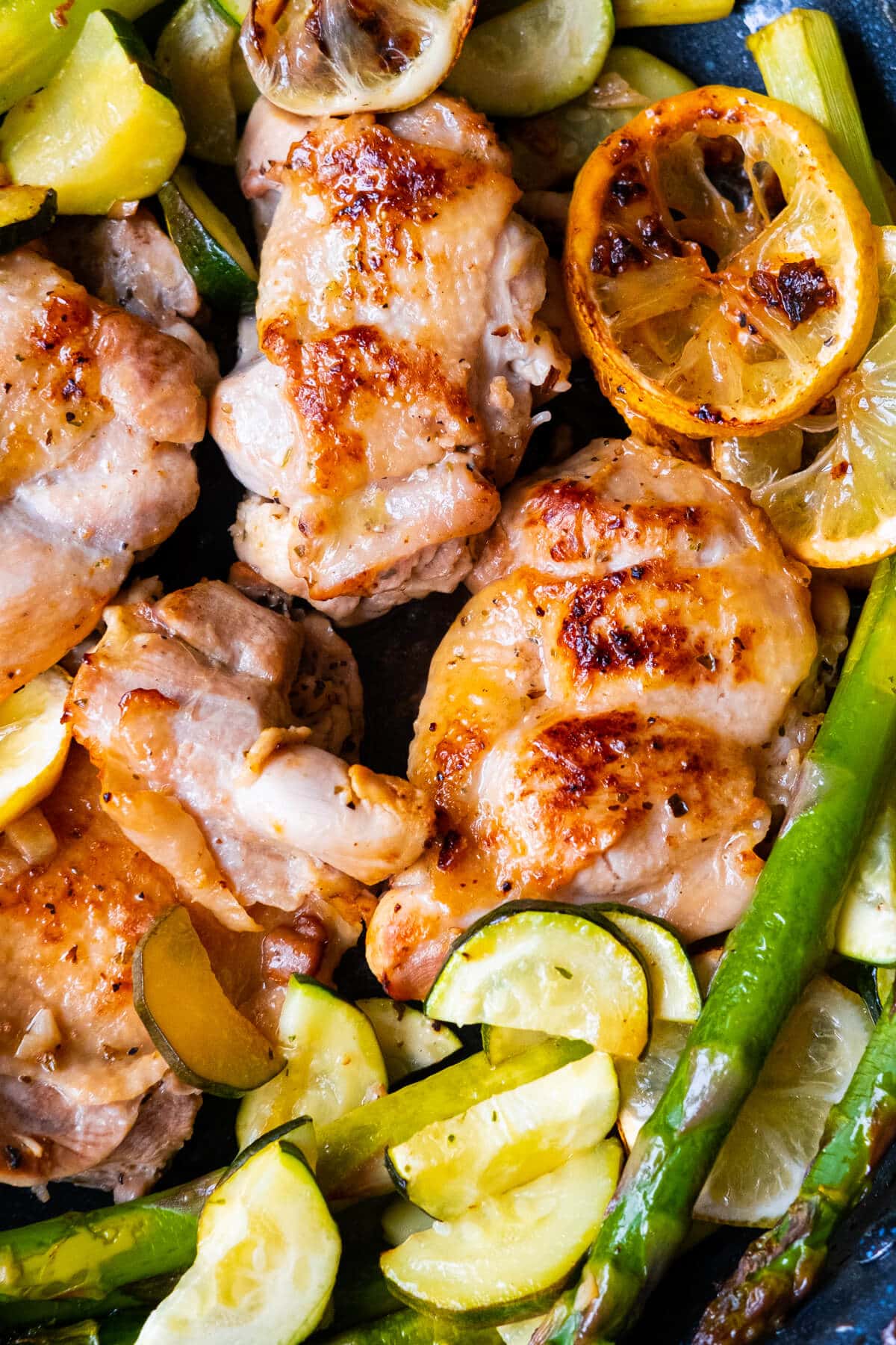 Golden brown seared chicken thighs alongside asparagus, zucchini slices and lemon slices. 