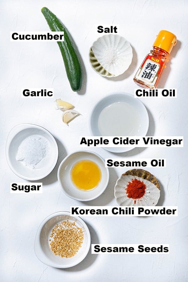 Asian cucumber salad ingredients photo showing raw cucumber, salt, chili oil, vinegar, chili powder, sesame oil, sugar and sesame seeds. 