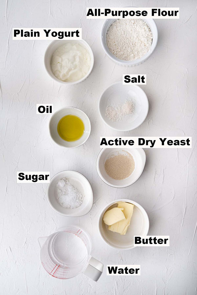 Picture of all the ingredients in naan recipe, close wise from the top: all-purpose flour, salt, active dry yeast, butter, water, sugar, oil and plain yogurt. 