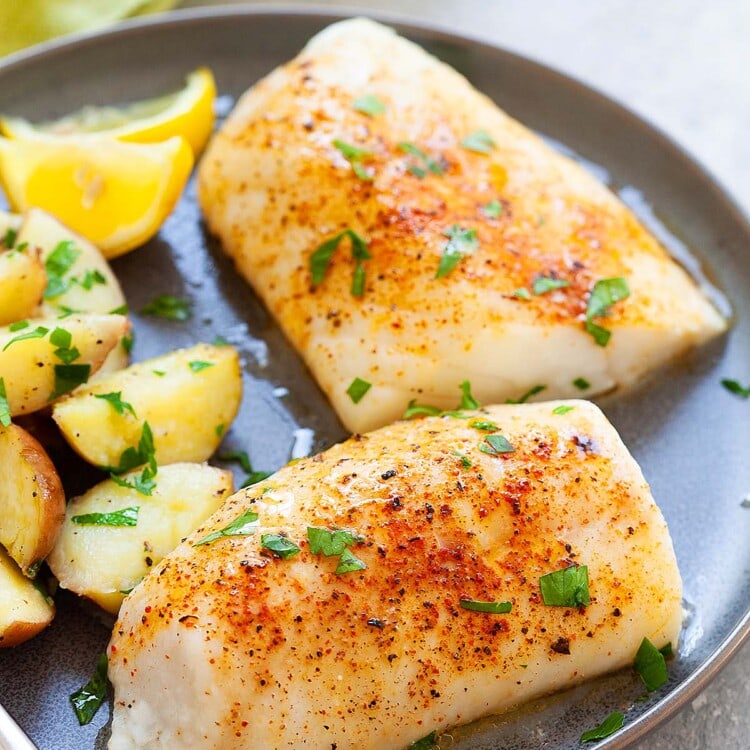 Freshly baked cod fillet with seasonings crust on a plate, garnished with lemon slices and herbs.