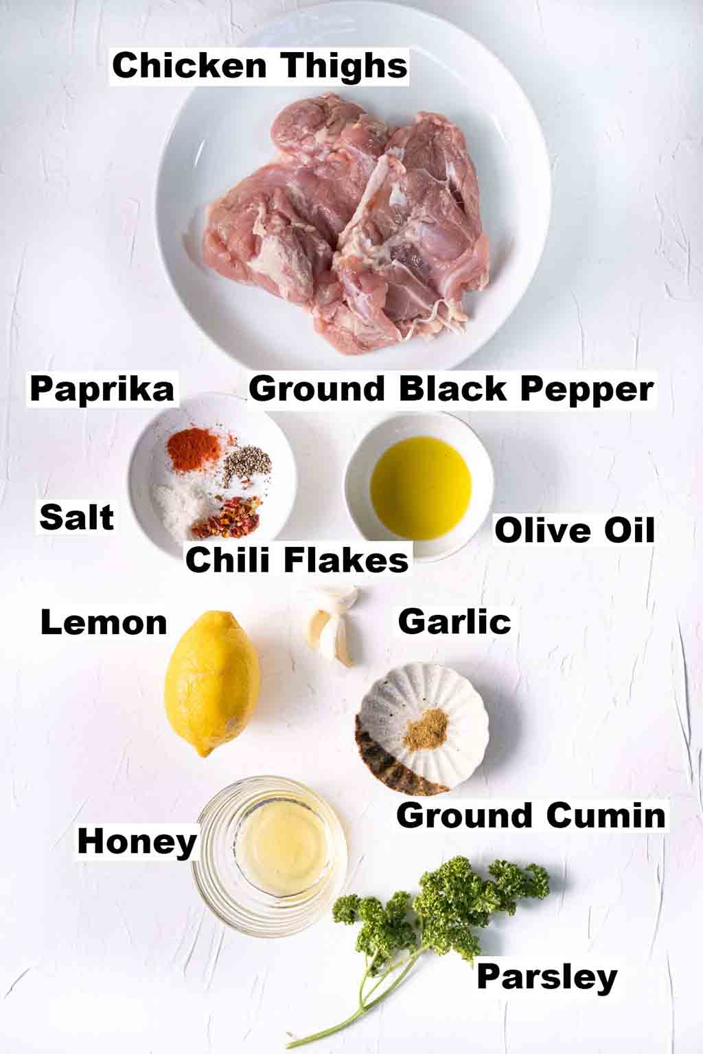 This image contains ingredients such as chicken thighs, paprika, ground black pepper, salt, chili flakes, olive oil. lemon, garlic, honey ground cumin and parsley which are needed to make the Lemon Garlic Chicken recipe.