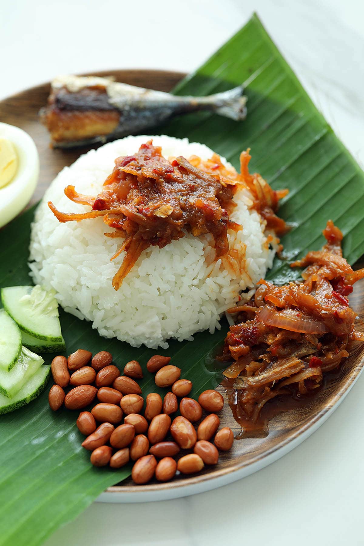 Nasi lemak on a banana leaf with sambal, anchovies, toasted peanuts, fried fish, and cucumber.