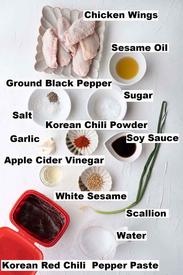 This image consists of ingredients such as chicken wings, sesame oil, ground black pepper, sugar, salt, Korean chili powder, garlic, soy sauce, apple cider vinegar, white sesame, scallion and Korean red chili pepper paste which are needed for the Spicy Korean Chicken Wings recipe.