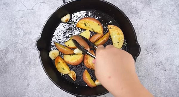 Potatoes being flipped with a tong in a cast-iron skillet with olive oil, butter and garlic. 