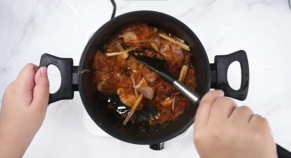 Spice paste, cinnamon, cloves, star anise, cardamom pods, lemongrass and chicken in a skillet being stirred with a spatula. 