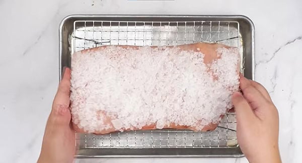 Pork belly covered in kosher salt is placed on a wire rack on top of a baking tray with water. 