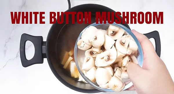 White button mushrooms being poured into a pot with soft tofu and chicken broth. 
