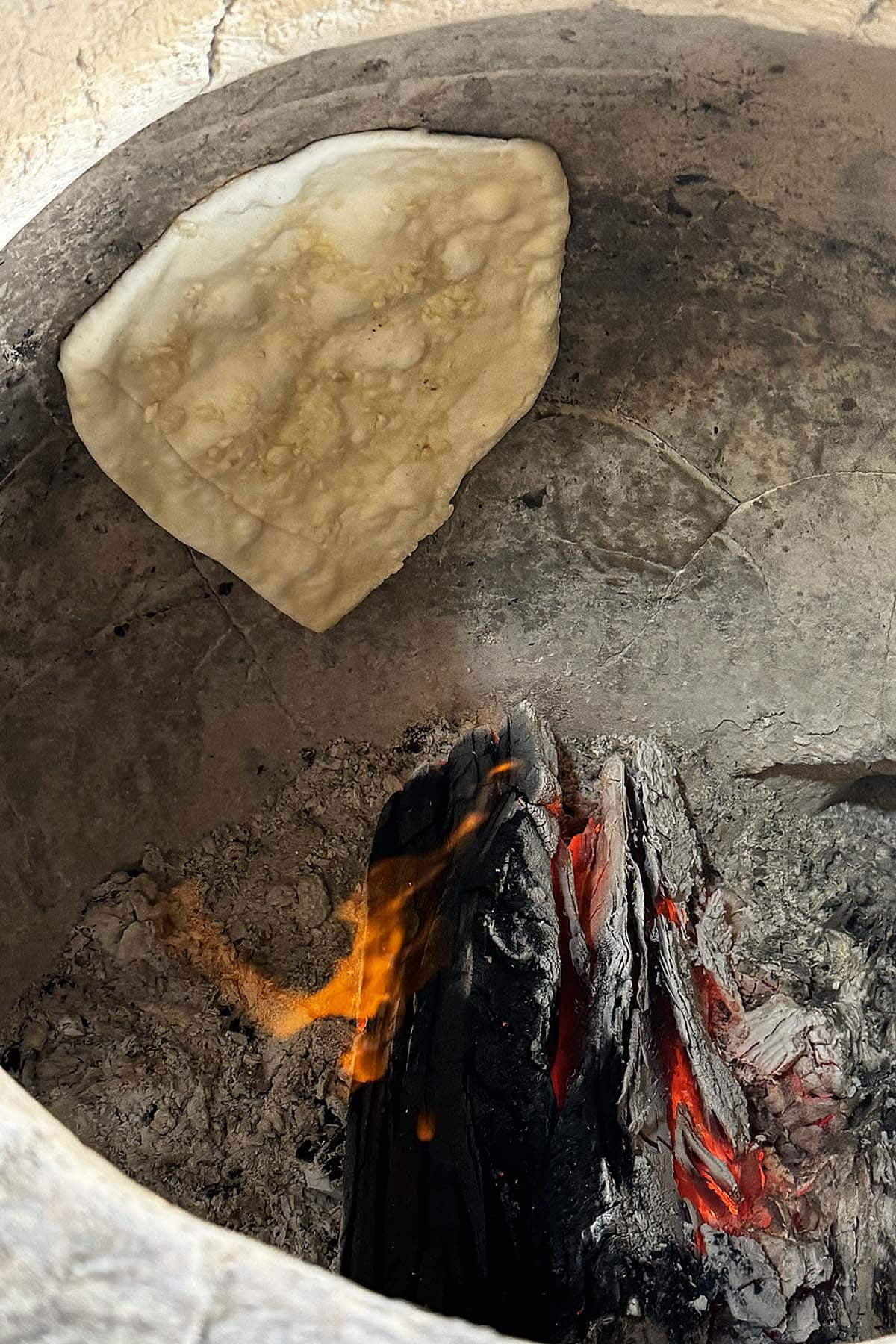 A piece of naan bread bubbles up in a traditional clay tandoor oven, burning with charcoal fire.