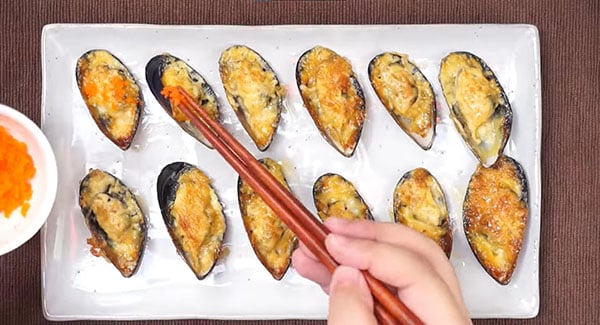 Cheese-mayo baked mussels being topped with tobiko fish roes using a pair of chopsticks. 