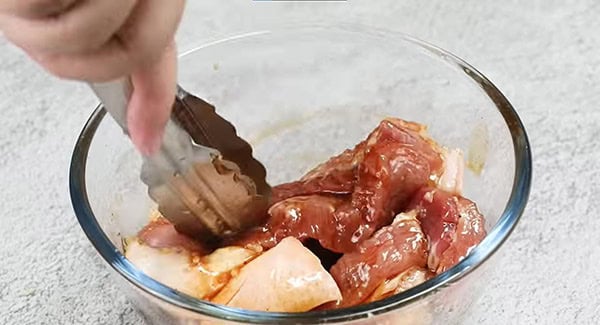 Pork belly and Chinese BBQ pork sauce are being mixed in a bowl with a pair of tongs.