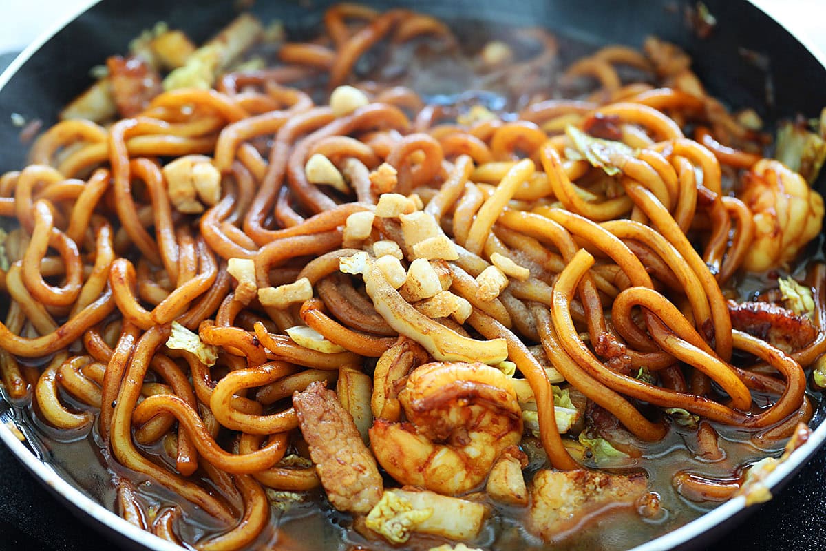 Cooking Malaysian hokkien mee with dark sauce in a skillet.