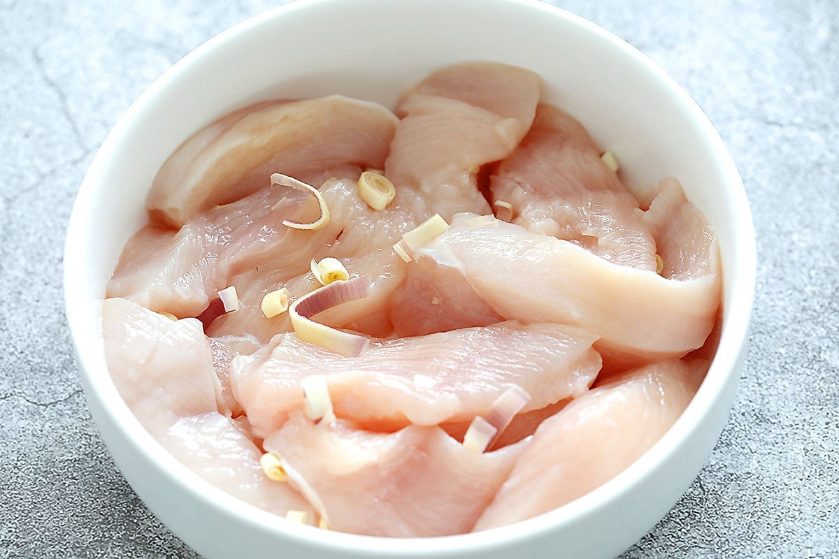 Raw chicken breast strips with fresh lemongrass slices in a bowl.