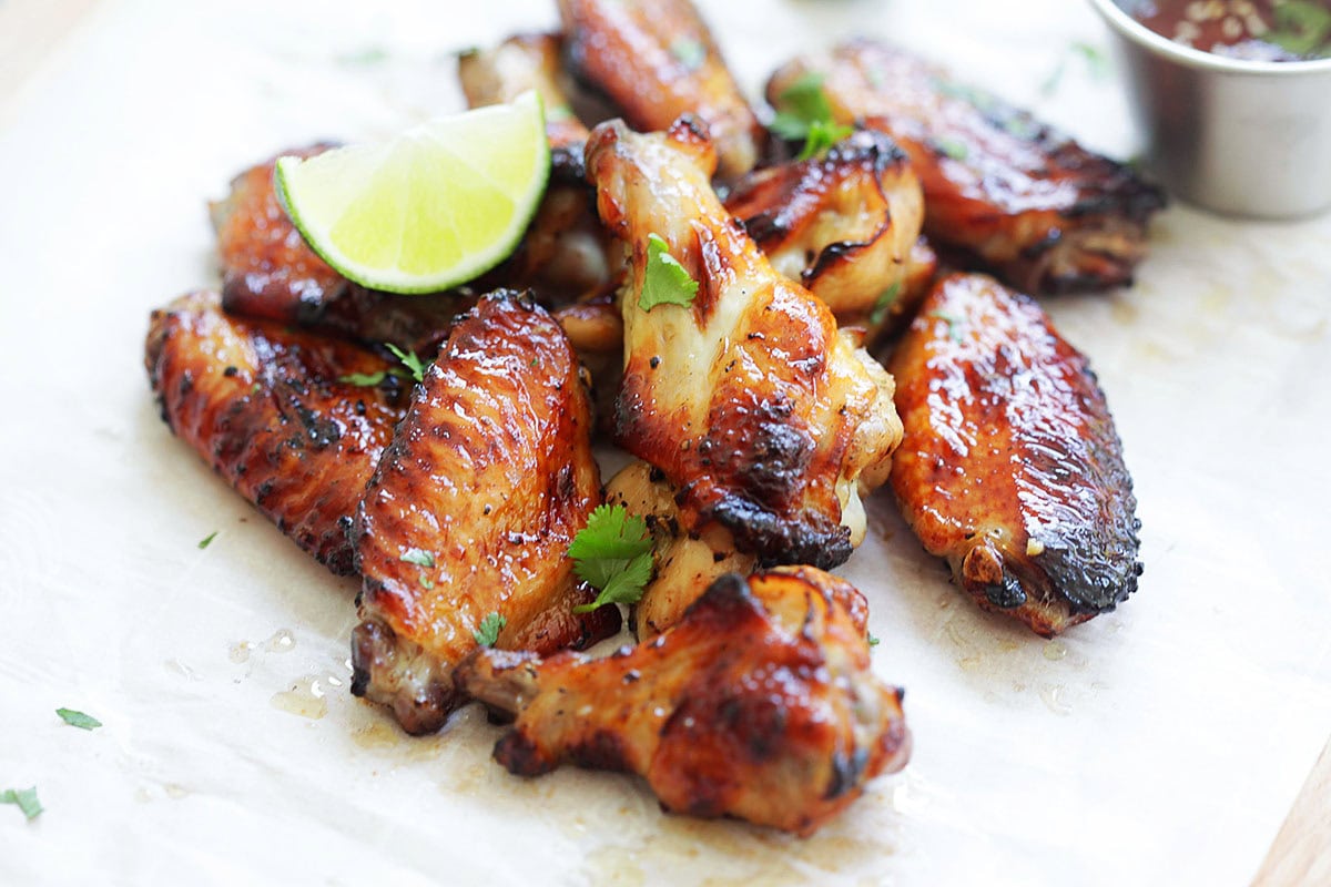 Thai grilled chicken wings with sweet chili sauce served on parchment paper, garnished with parsley, and paired with lime wedges and dipping sauce.