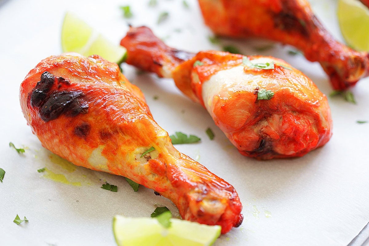Juicy tandoori chicken leg pieces with a vibrant red color, arranged on parchment paper with lime.