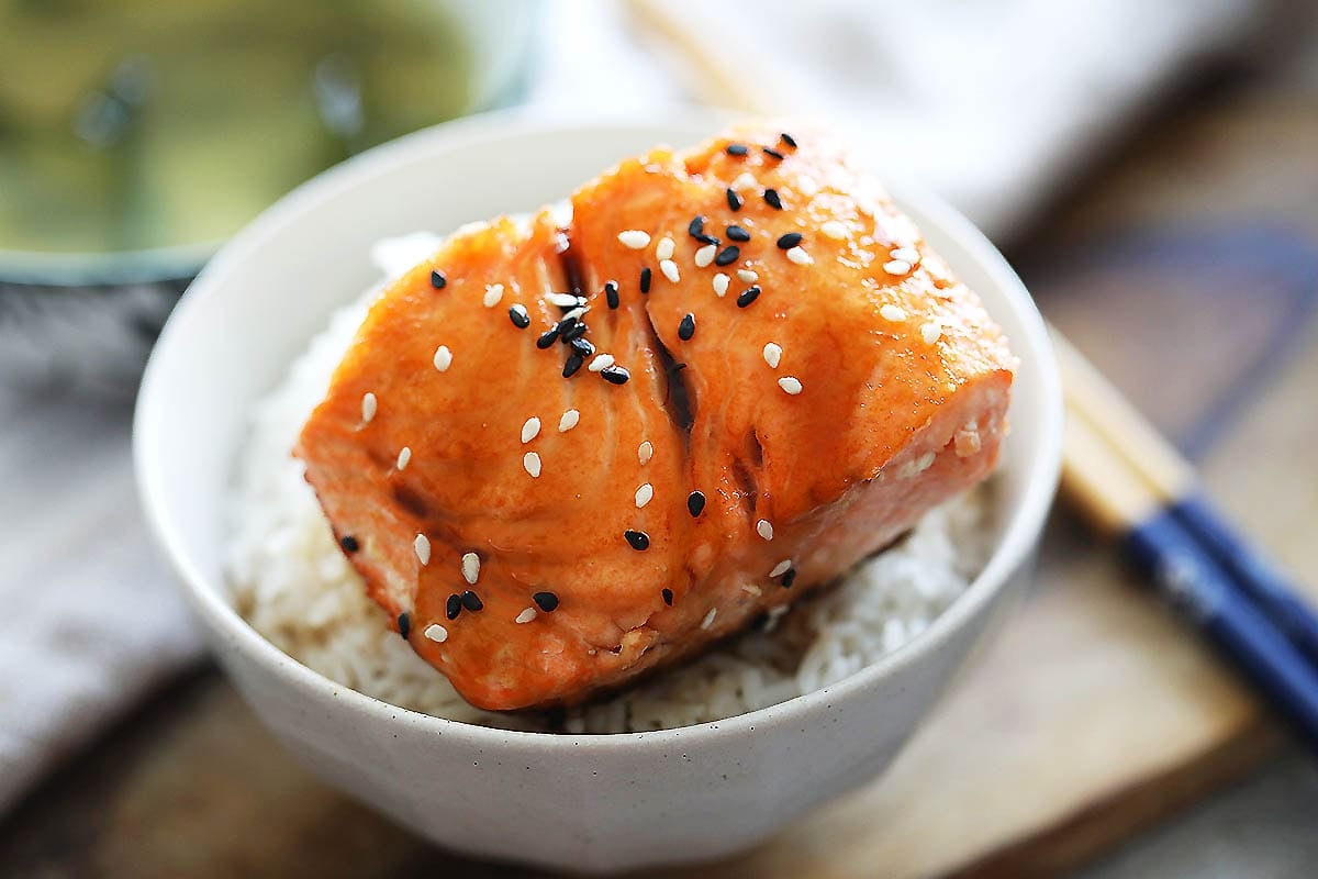 Salmon teriyaki topped with black and white sesame seeds, served on a bowl of rice.