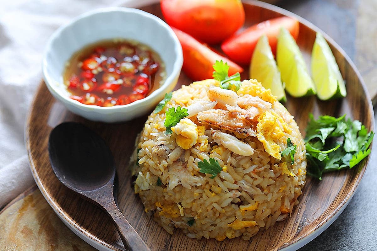 Crab fried rice on a plate with lime wedges, tomato wedges, cilantro, and nam pla prik.
